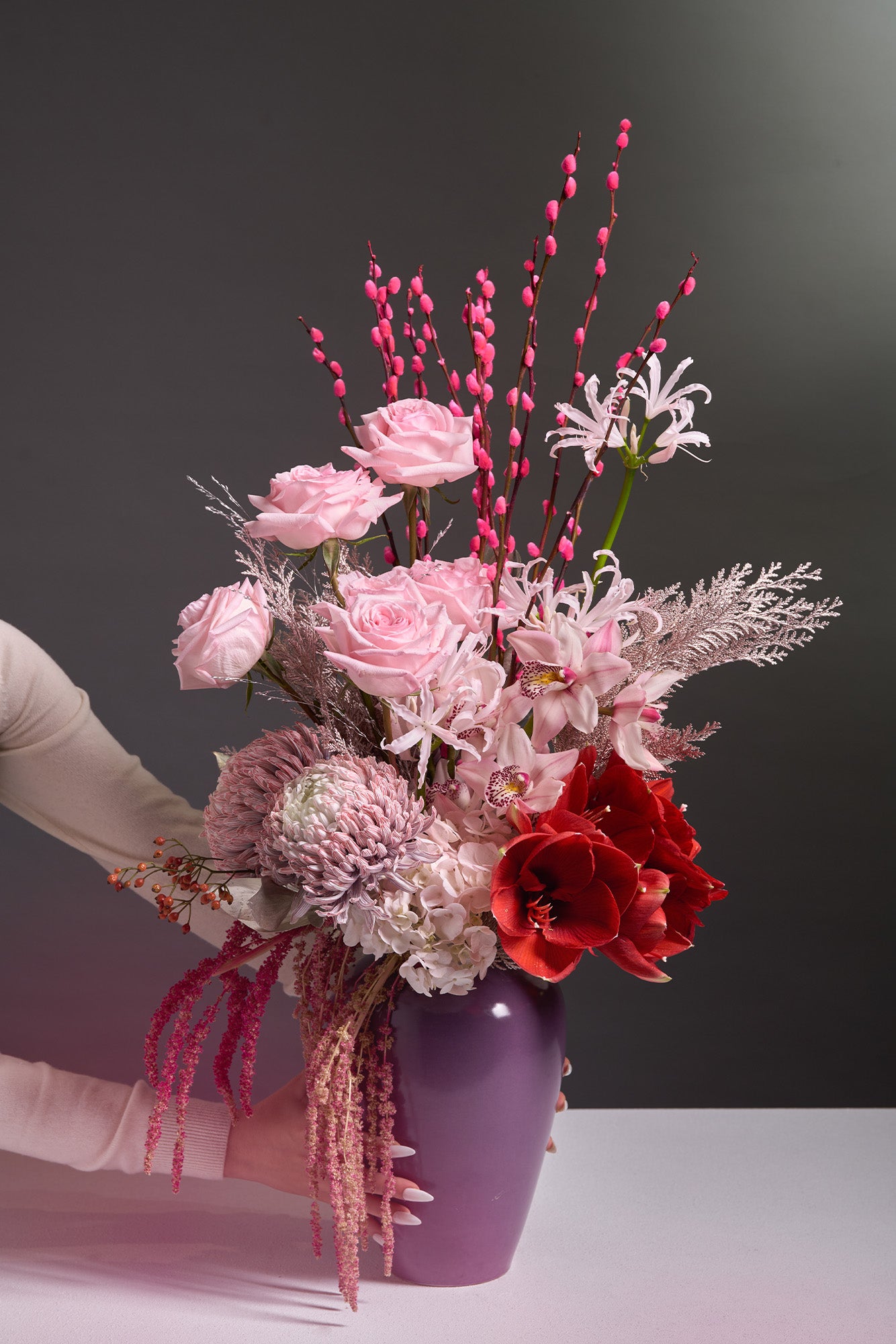 loral Elegance arrangement with pink roses, cymbidium orchids, lavender chrysanthemums, red amaryllis, pink pussy willows, and a bold purple vase, symbolising harmony and new beginnings.